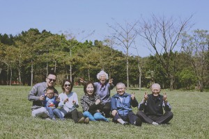 三代全家福,昭穎、文妍與兒子、孩子的爺爺奶奶和外公外婆坐在台北大學草地上,面向鏡頭笑著比手勢。